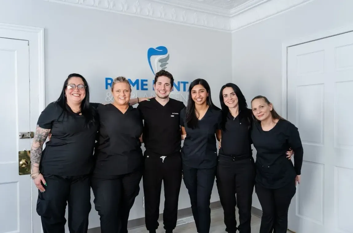 Six people in black outfits pose in a bright office with a blue logo on the wall.