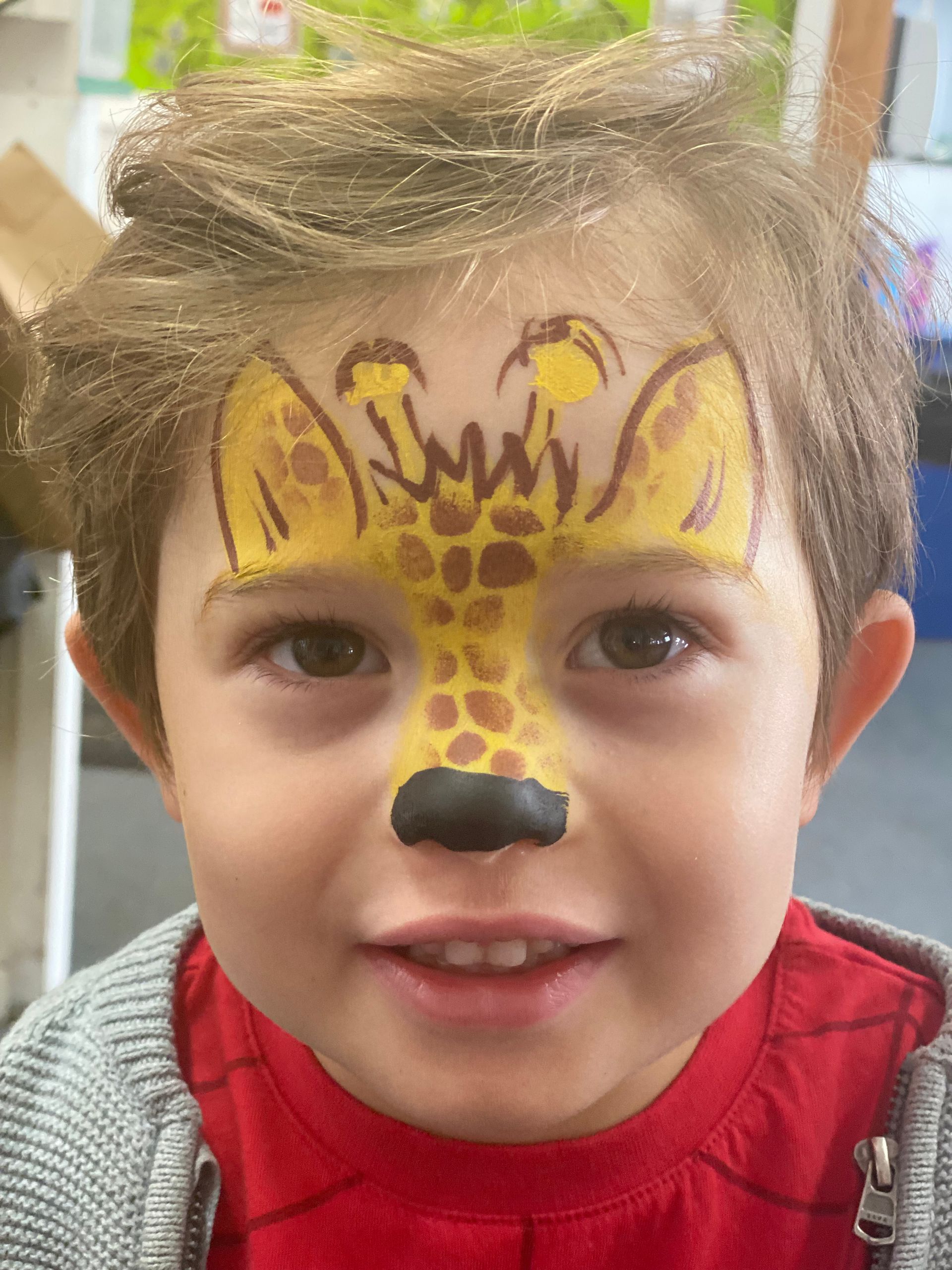 Boy with giraffe face paint: yellow and brown spots, black nose, smiling.