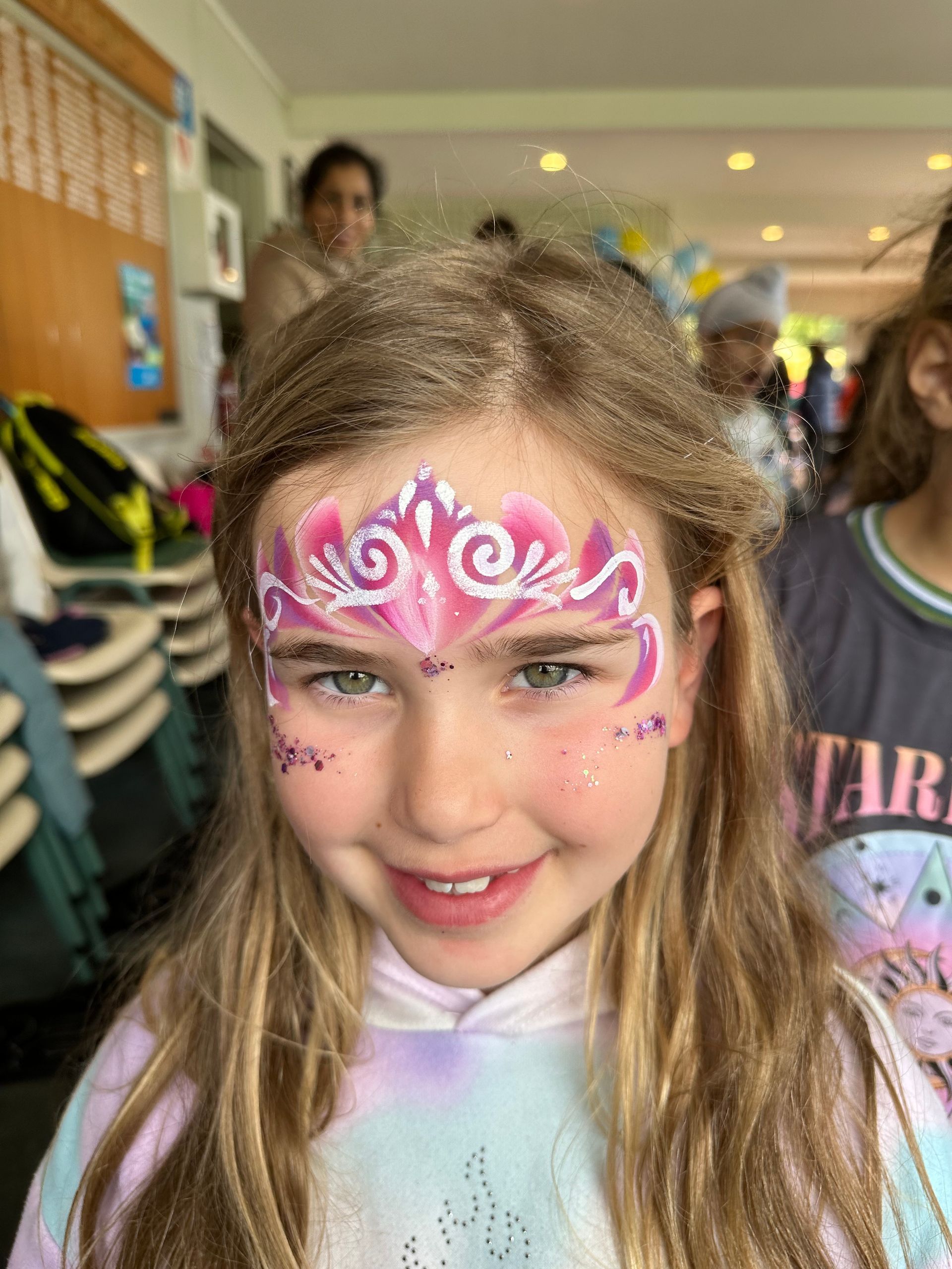 Girl with pink and purple face paint smiling; outdoors.