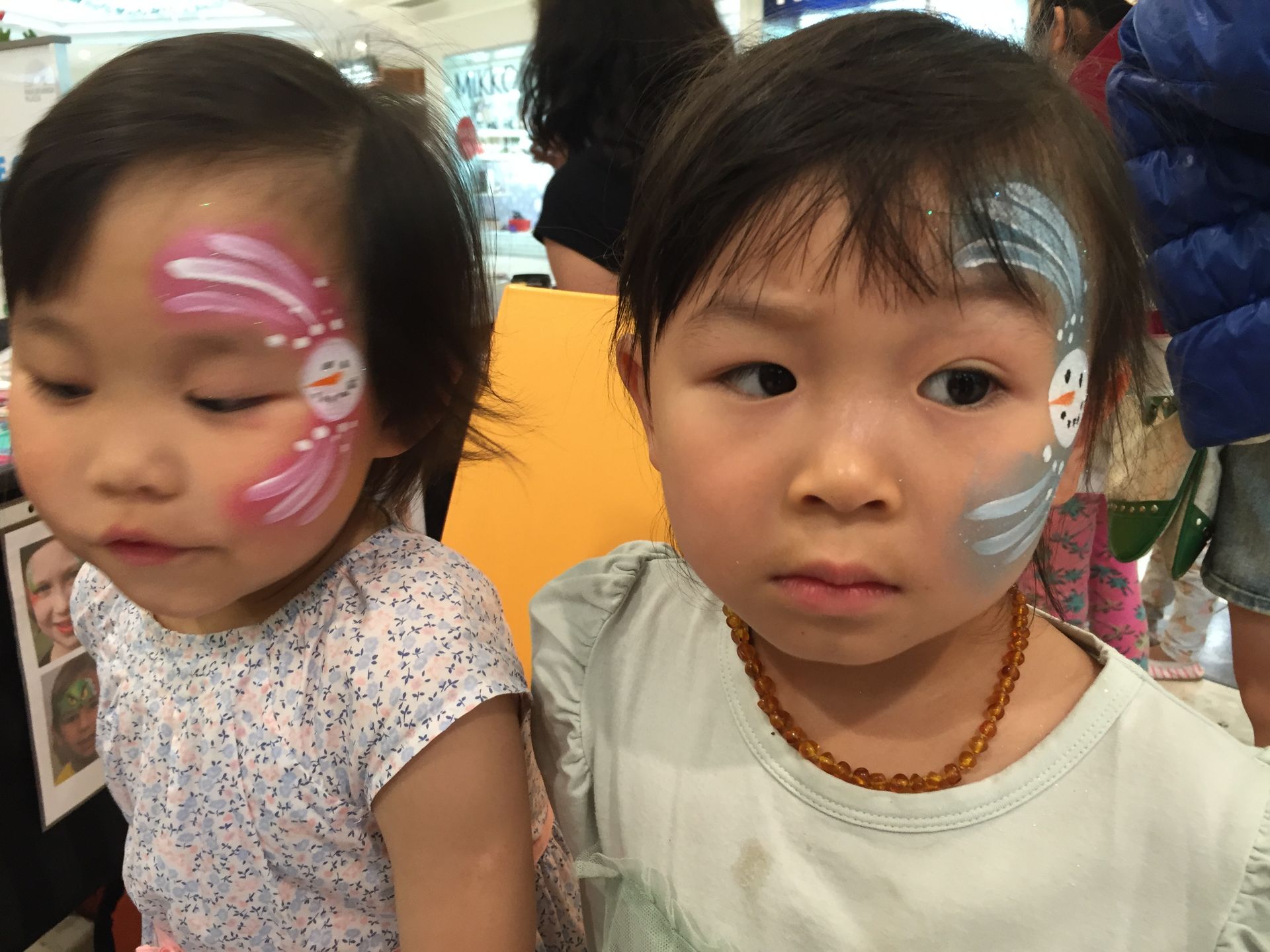 Two young Asian girls with face paint, possibly at an event. One has a snowman design; the other, a flower design.