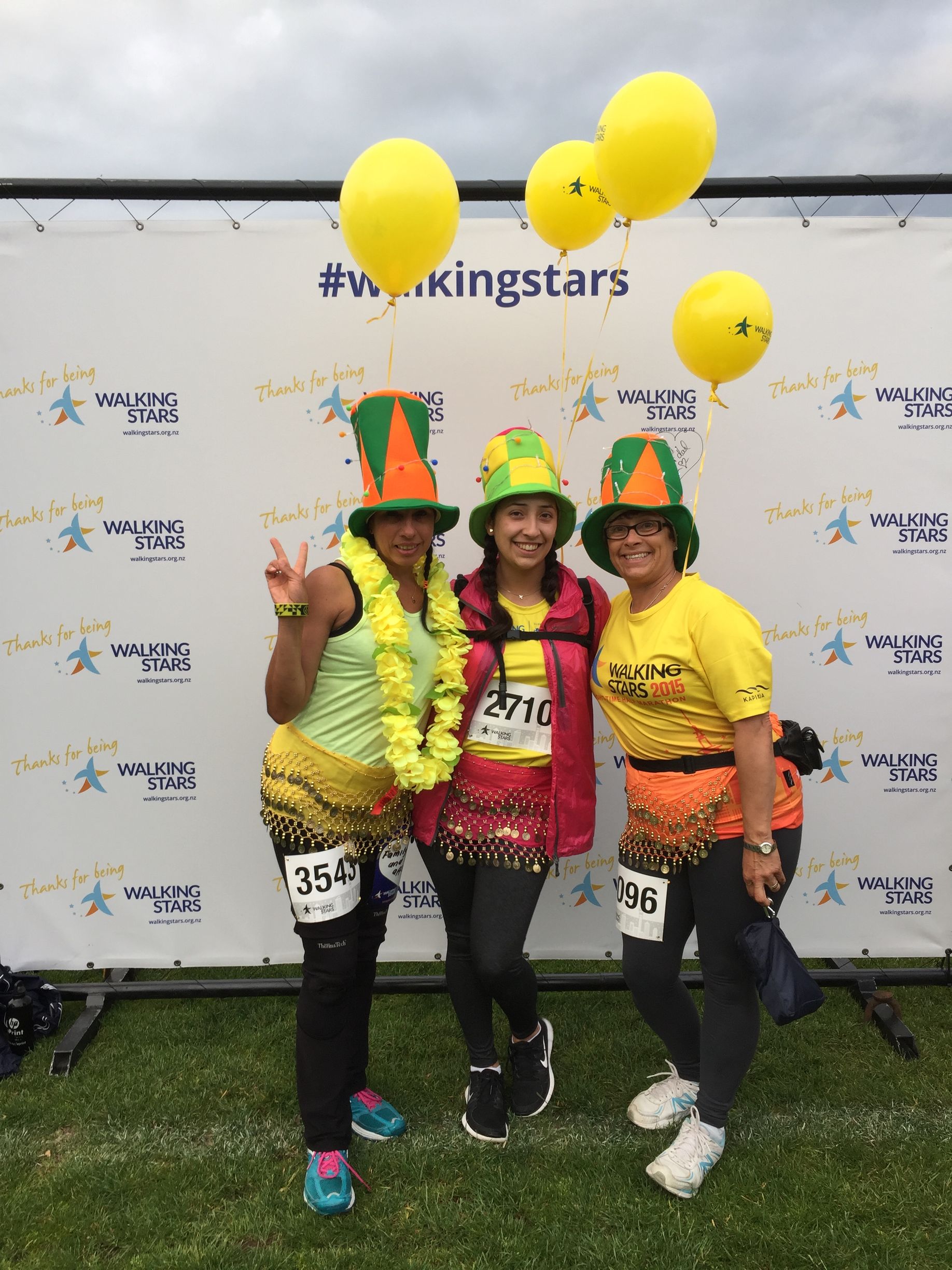 Three women in colorful costumes, posing in front of a backdrop with yellow balloons.