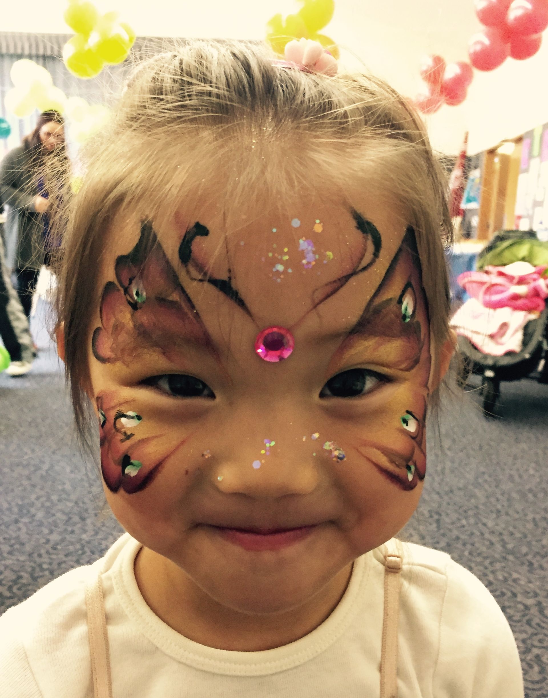Young girl with butterfly face paint smiles at the camera, decorated with glitter and a pink gem.