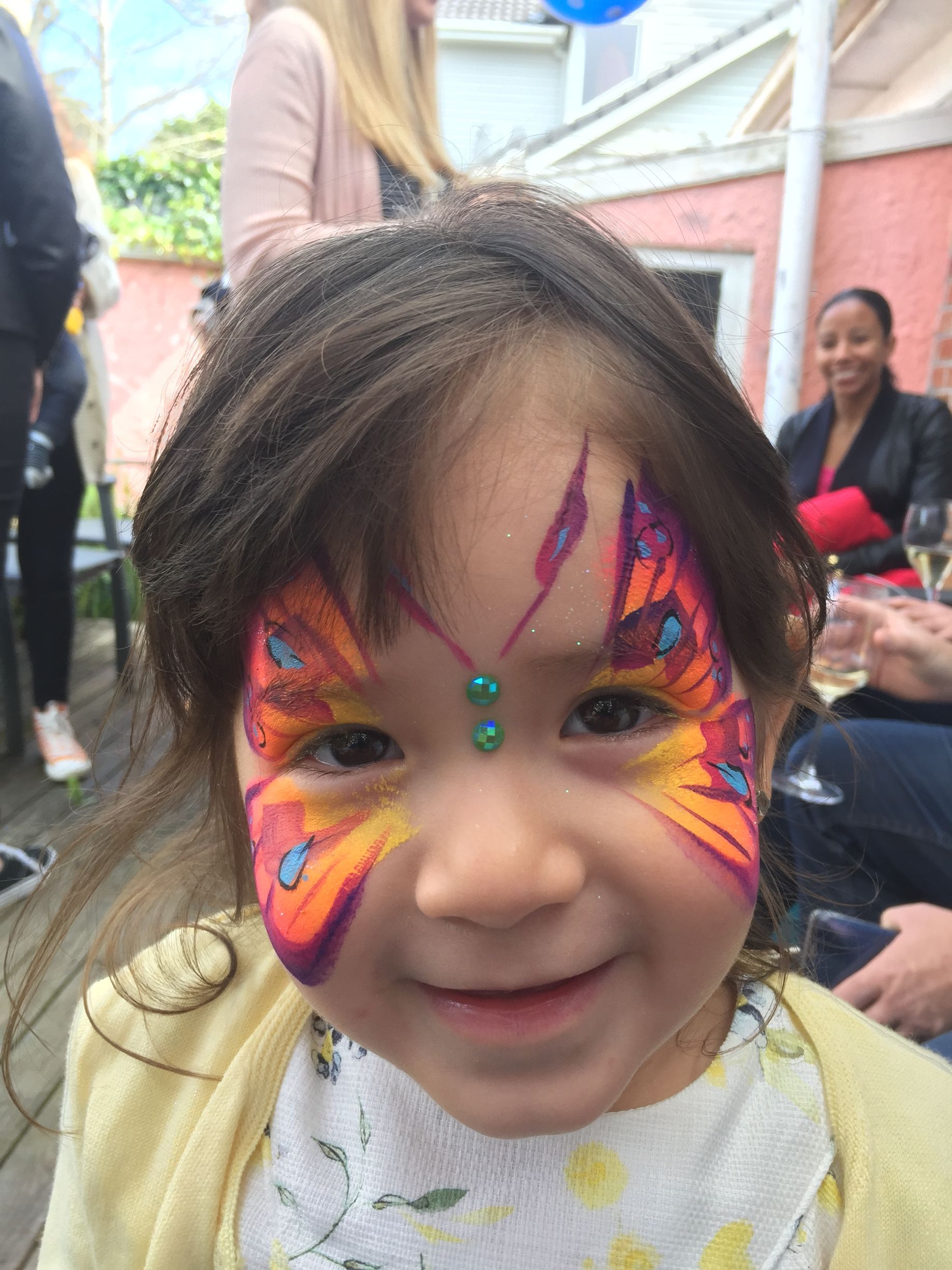 Girl with butterfly face paint smiles, enjoying an outdoor party.