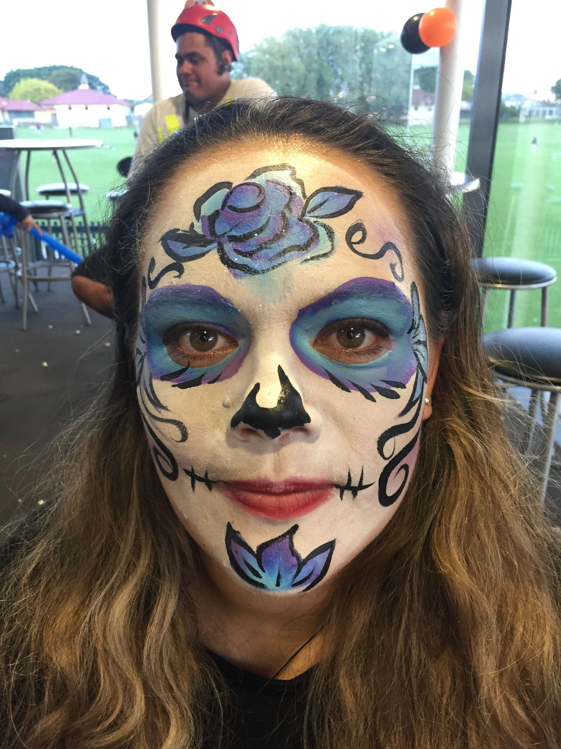 Woman with Day of the Dead face paint, featuring blue and black details.  Background includes a man and outdoor setting.