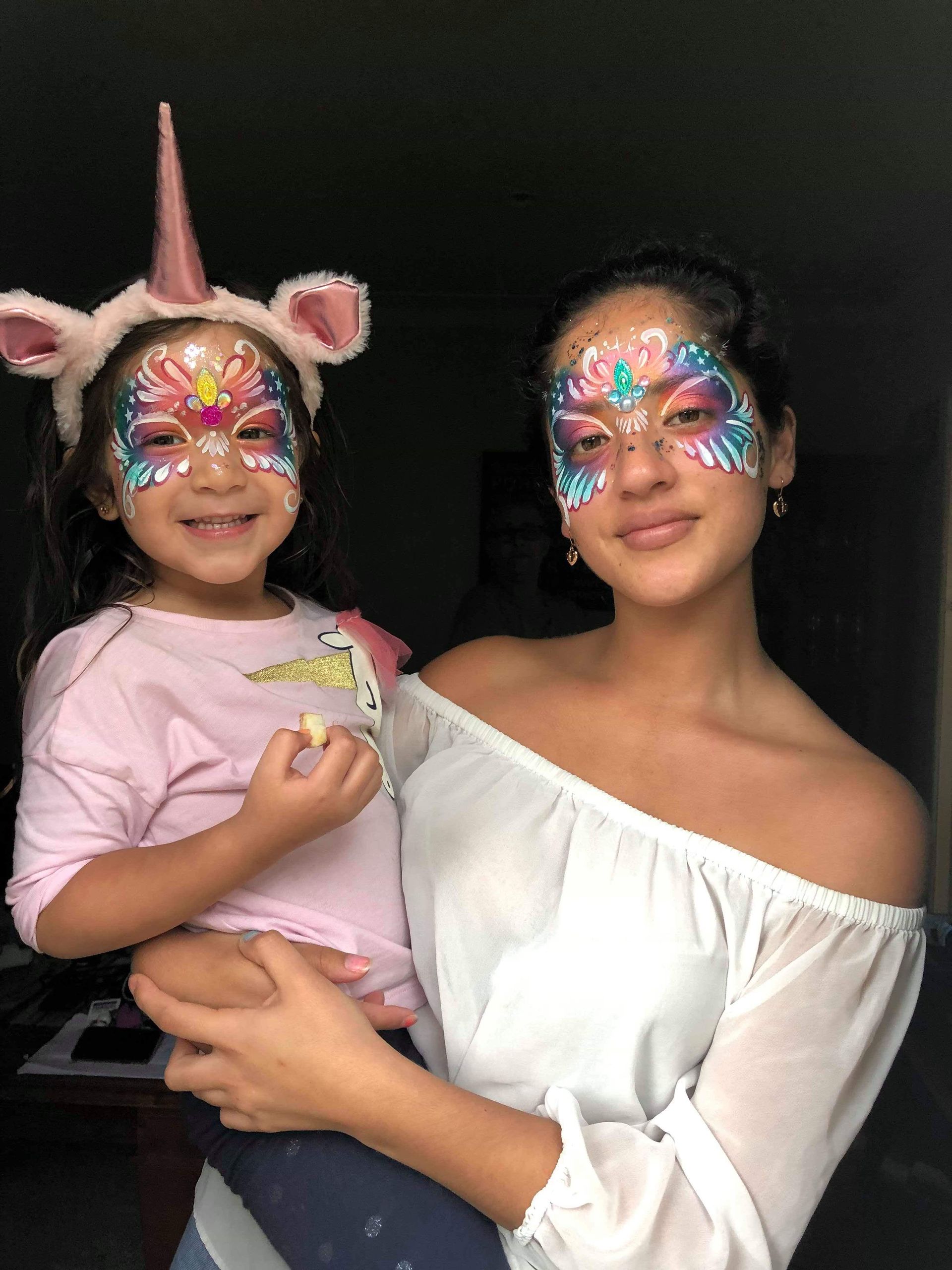 Woman holds a child; both have face paint and smiling. Child wears unicorn headband. Dark setting.