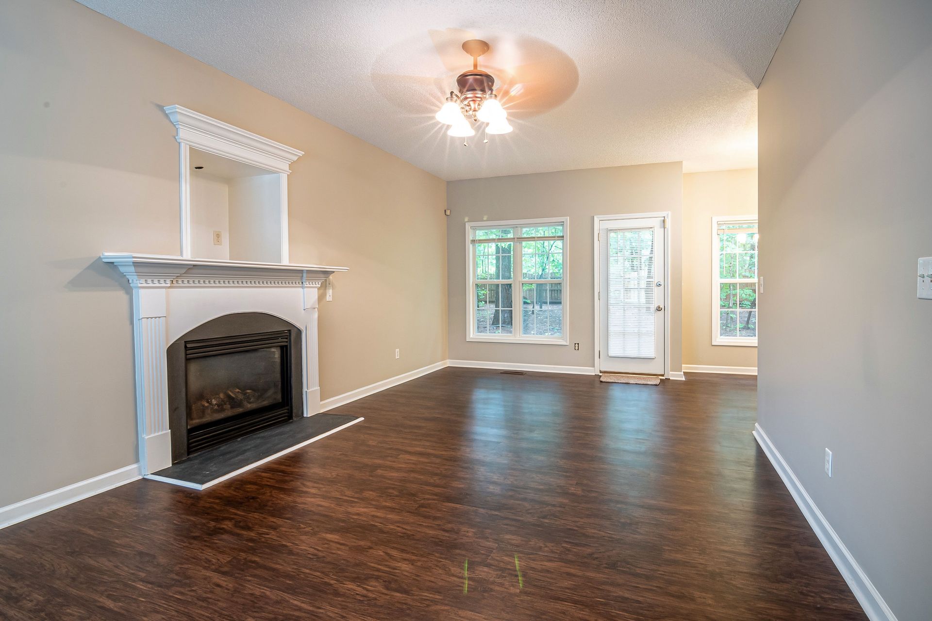 Empty living room with dark hardwood floors, white fireplace mantel, and large windows.