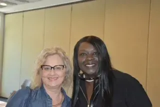 Two people smiling and posing together indoors against a beige wall.