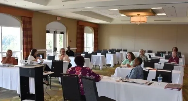 People seated at spaced tables in a bright conference room, some using laptops and papers