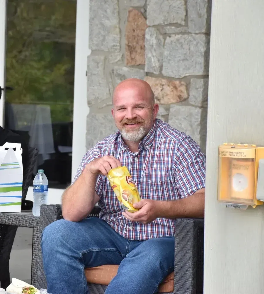Smiling man sitting on a porch holding a bag of chips, with a water bottle and stone wall behind him