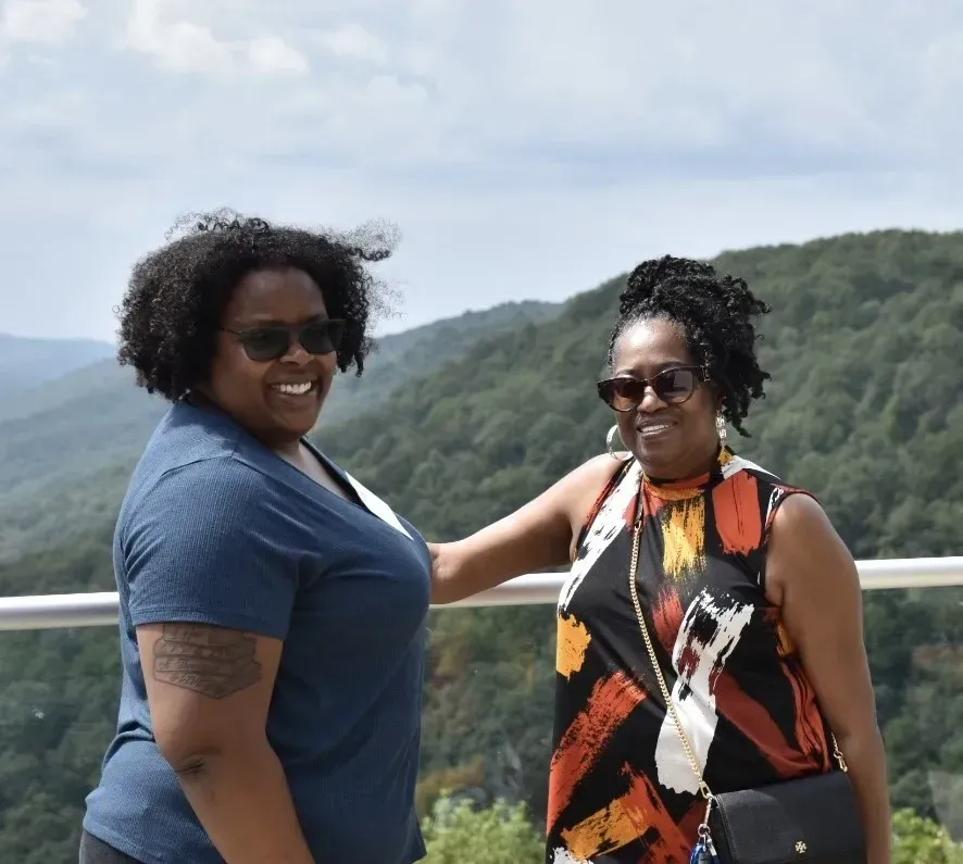 Two people smiling on a scenic overlook with forested mountains in the background.