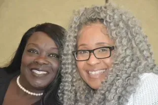 Two smiling people posing close together against a beige background, one in black and pearl necklace, one with gray curls and glasses