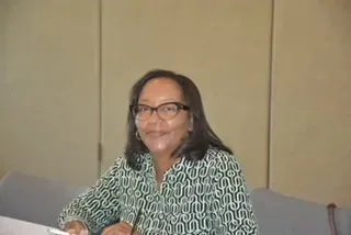 Woman smiling at a table in a beige room, wearing glasses and a green patterned shirt.
