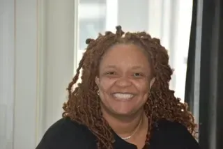 Smiling person with shoulder-length curly hair in a black top, standing indoors near a window.