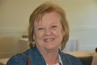 Smiling woman in a blue denim jacket standing indoors against a light wall