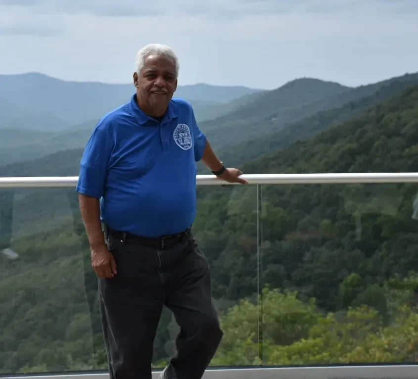 Man in blue polo standing by a railing with mountain views in the background.