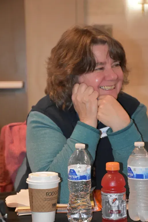 Smiling woman seated at a table with coffee cups and water bottles in a casual indoor setting