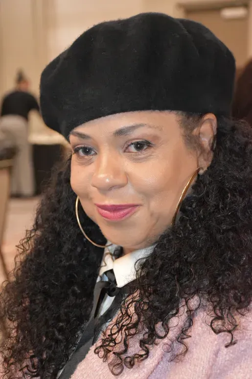 Smiling person in black beret and pink jacket, with curly hair, at an indoor event.