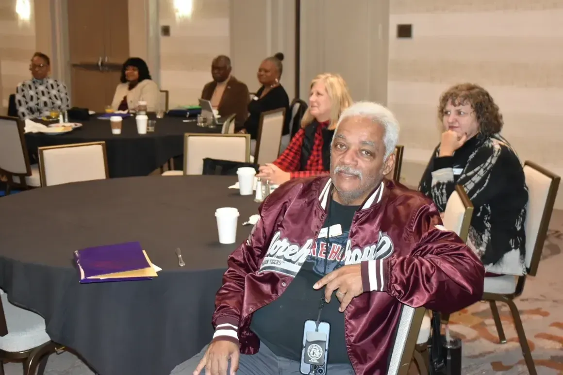 Conference attendees seated at tables in a meeting room, one man in a maroon jacket in the foreground