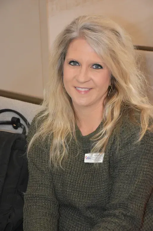Blonde woman smiling in a dark green sweater, seated indoors with a name badge.