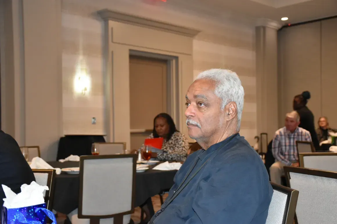 Man in a blue shirt seated at a conference table in a hotel meeting room, looking to the side