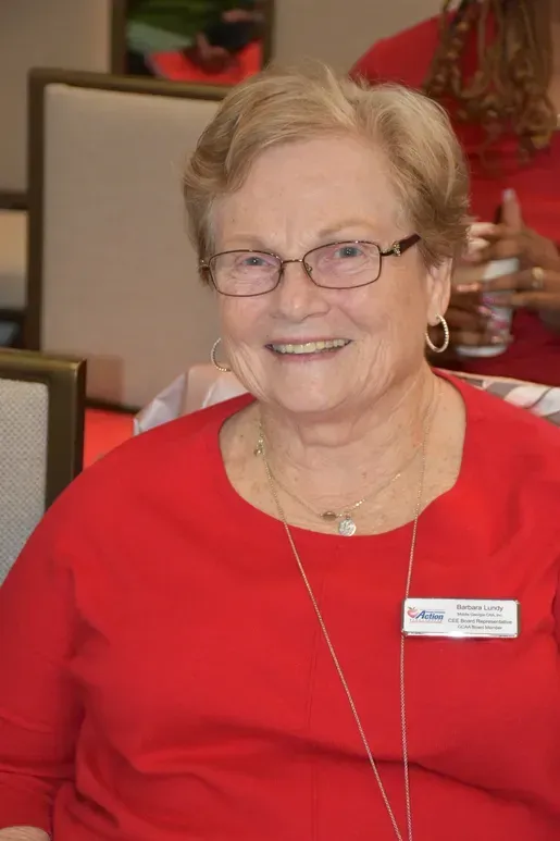 Smiling woman in red top with glasses and name badge, seated indoors at an event