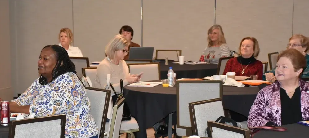 People seated at tables in a meeting room, listening and taking notes