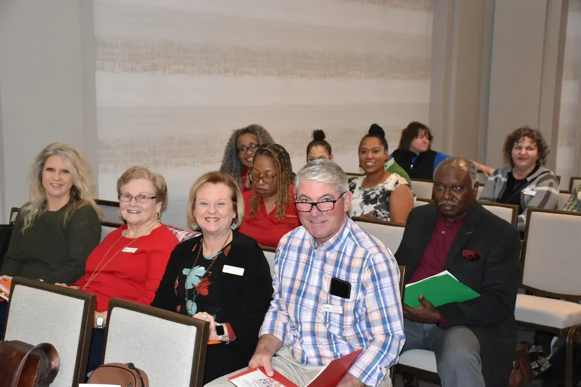 Audience seated in a conference room, holding folders and looking toward the front.