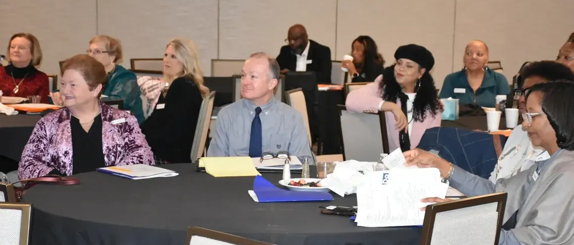 People seated at tables in a meeting room, listening and taking notes.