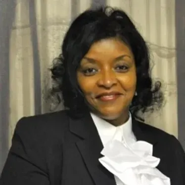 Portrait of a smiling woman in a black blazer with a white ruffled blouse, standing before curtains