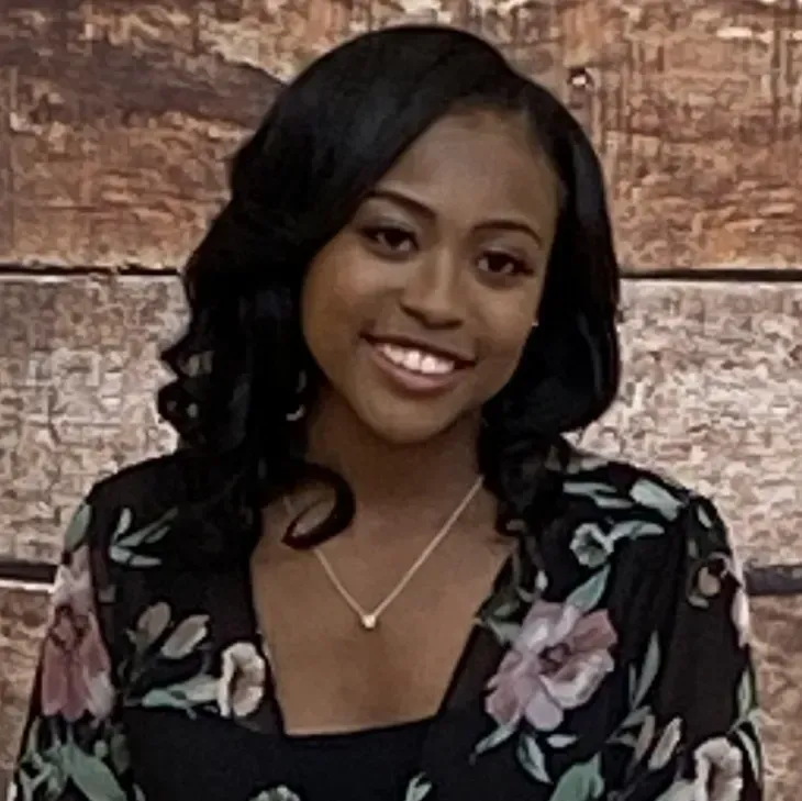 Smiling woman in a black floral top and necklace against a brick wall background
