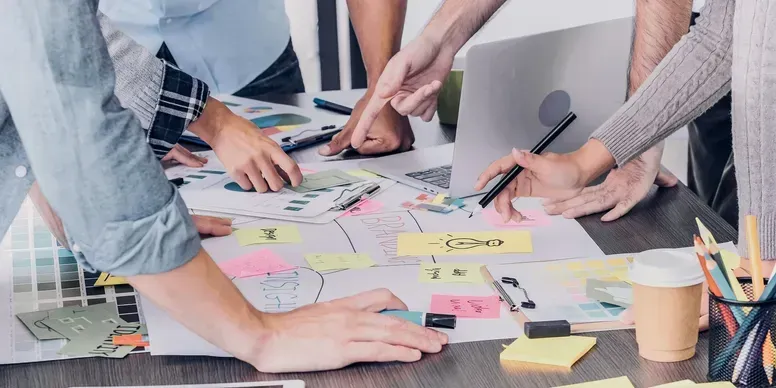 Hands collaborating over a table covered with sticky notes, papers, and a coffee cup.