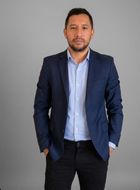 Persona sonriente con un blazer oscuro y una blusa blanca sobre un fondo gris.