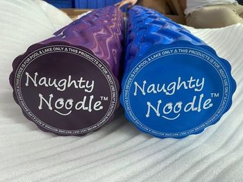 Two Naughty Noodle novelty tubes, one purple and one blue, on a white bedspread