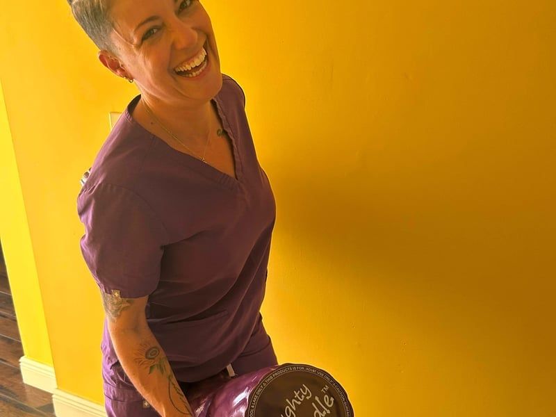 Person in purple scrubs smiling against a bright yellow wall, holding a round brown object.