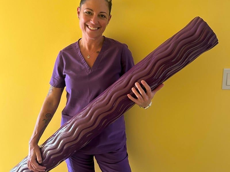 Woman in purple scrubs holding a large purple yoga mat against a yellow wall