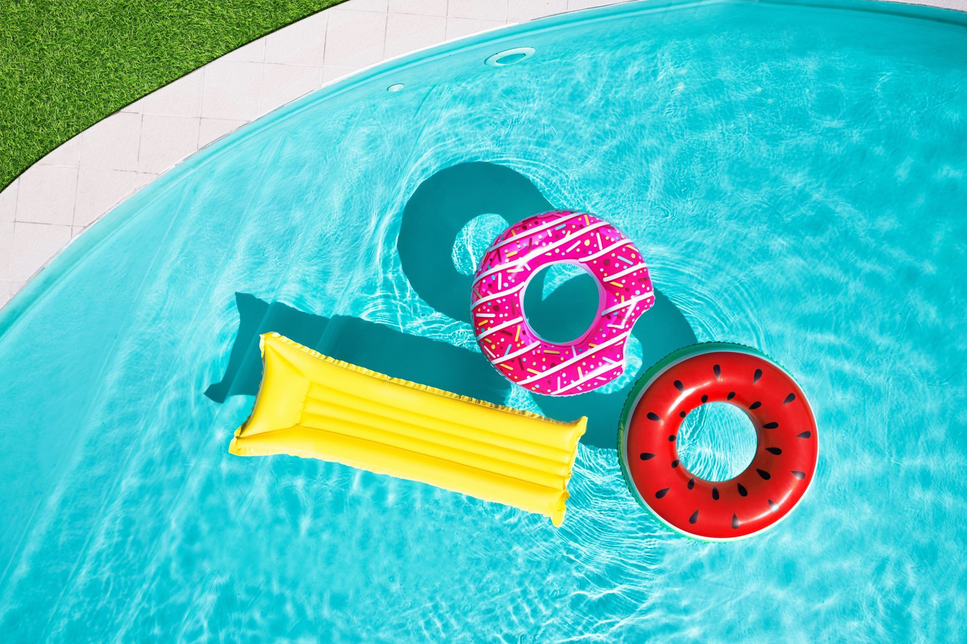 Three colorful pool floats in a swimming pool: yellow lounger, pink ring, and red ring with sunglasses