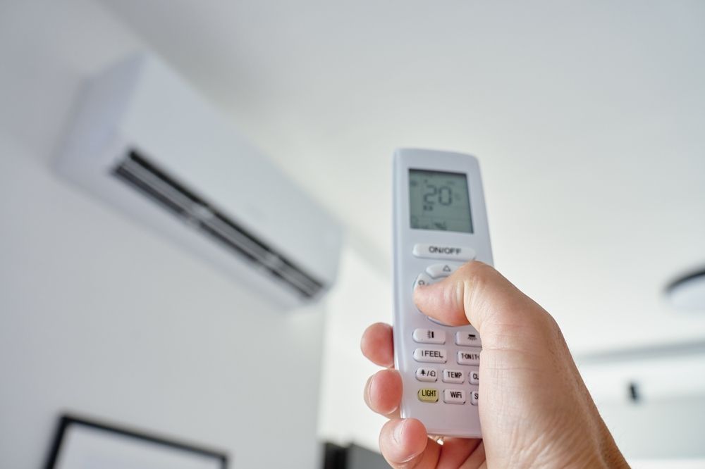 Hand holding a remote pointed at a wall-mounted air conditioner in a bright room