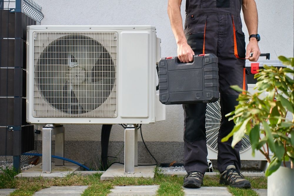 Technician standing beside an outdoor air conditioner unit, holding a black toolbox in a yard