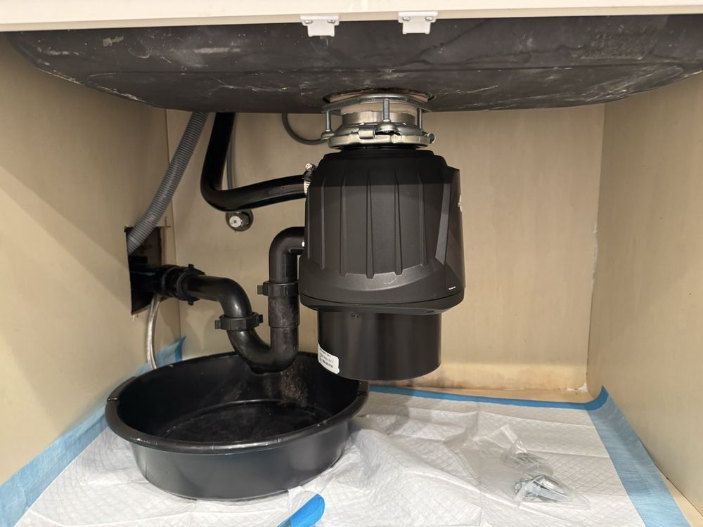 Under-sink cabinet with garbage disposal, plumbing pipes, and a black drain pan on a protective mat.