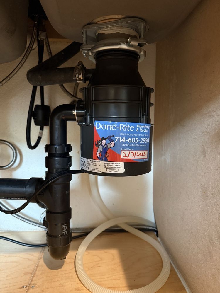 Garbage disposal under a sink with plumbing, black unit, and a clear tube looped below.