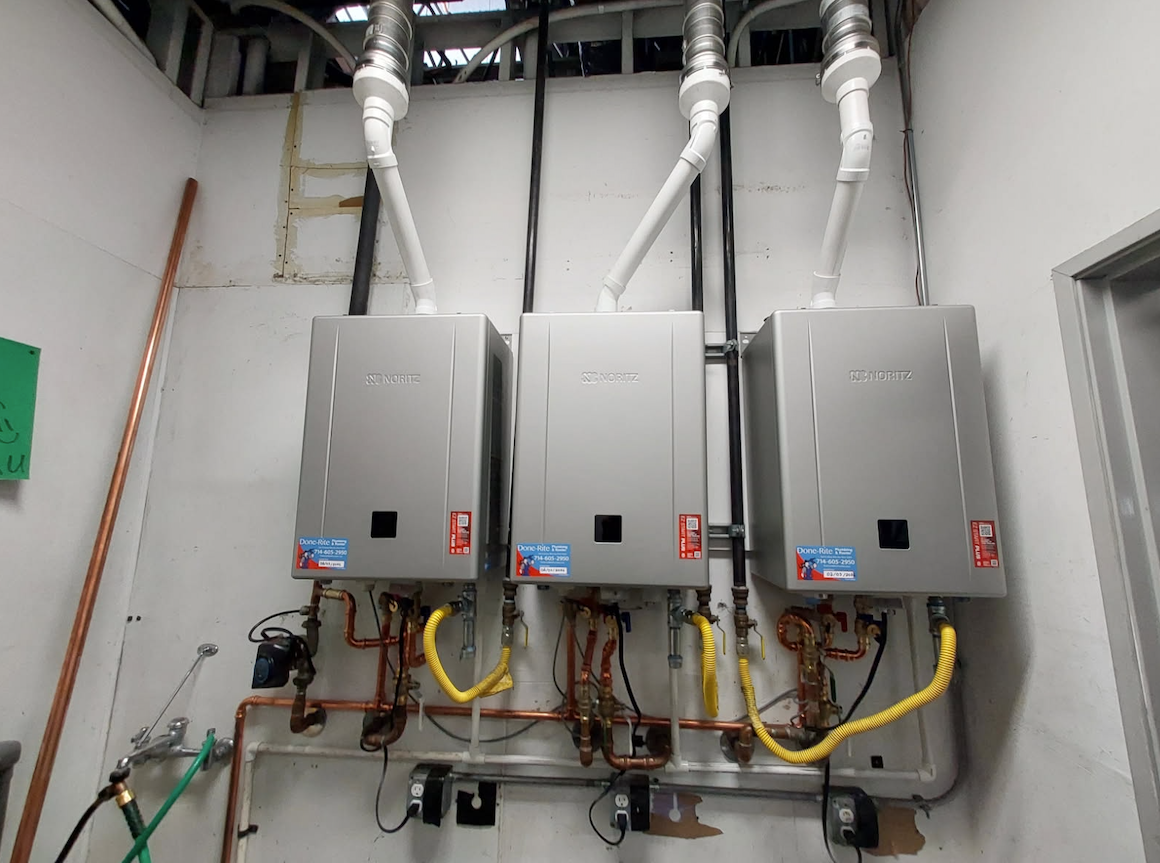 Three gray electrical panels with pipes and wiring mounted on a utility room wall