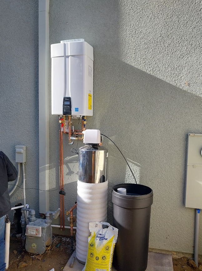 Outdoor water heater with insulated pipes beside a stucco wall and utility bins on the ground
