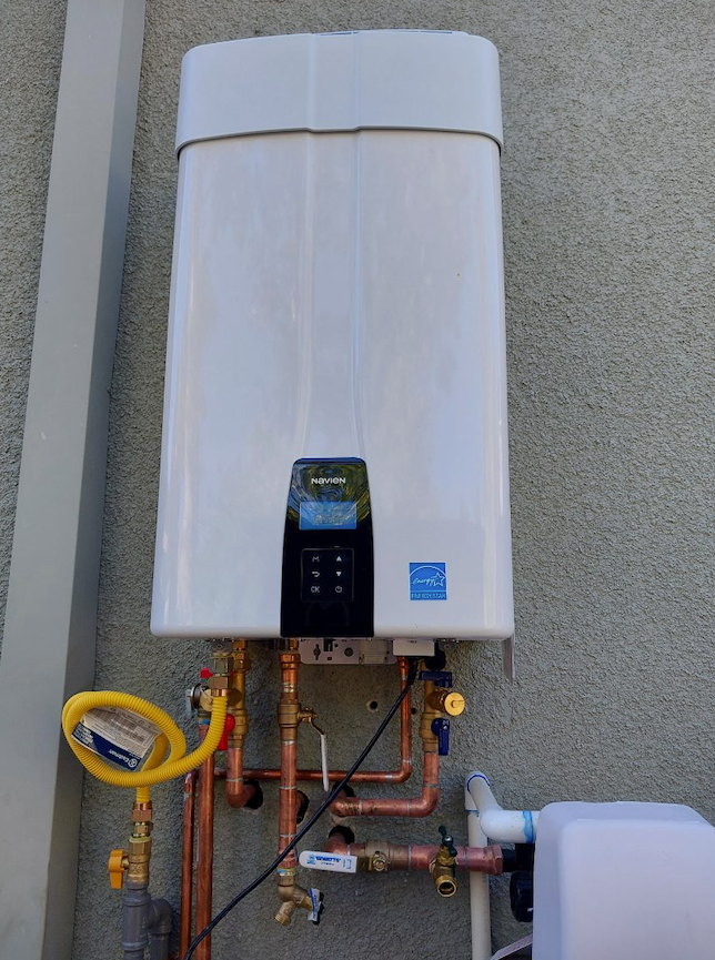 White wall-mounted tankless water heater with exposed pipes and hoses on an exterior wall.