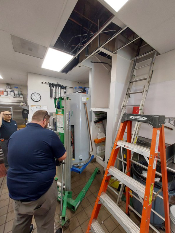 Workers repairing a ceiling opening in an office, with ladders and a lift under exposed framing.