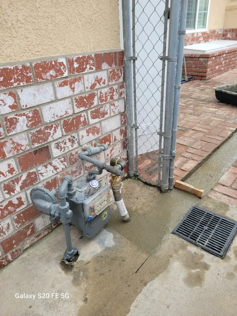 Gas meter beside a chain-link gate on a concrete sidewalk near a brick wall and drain grate