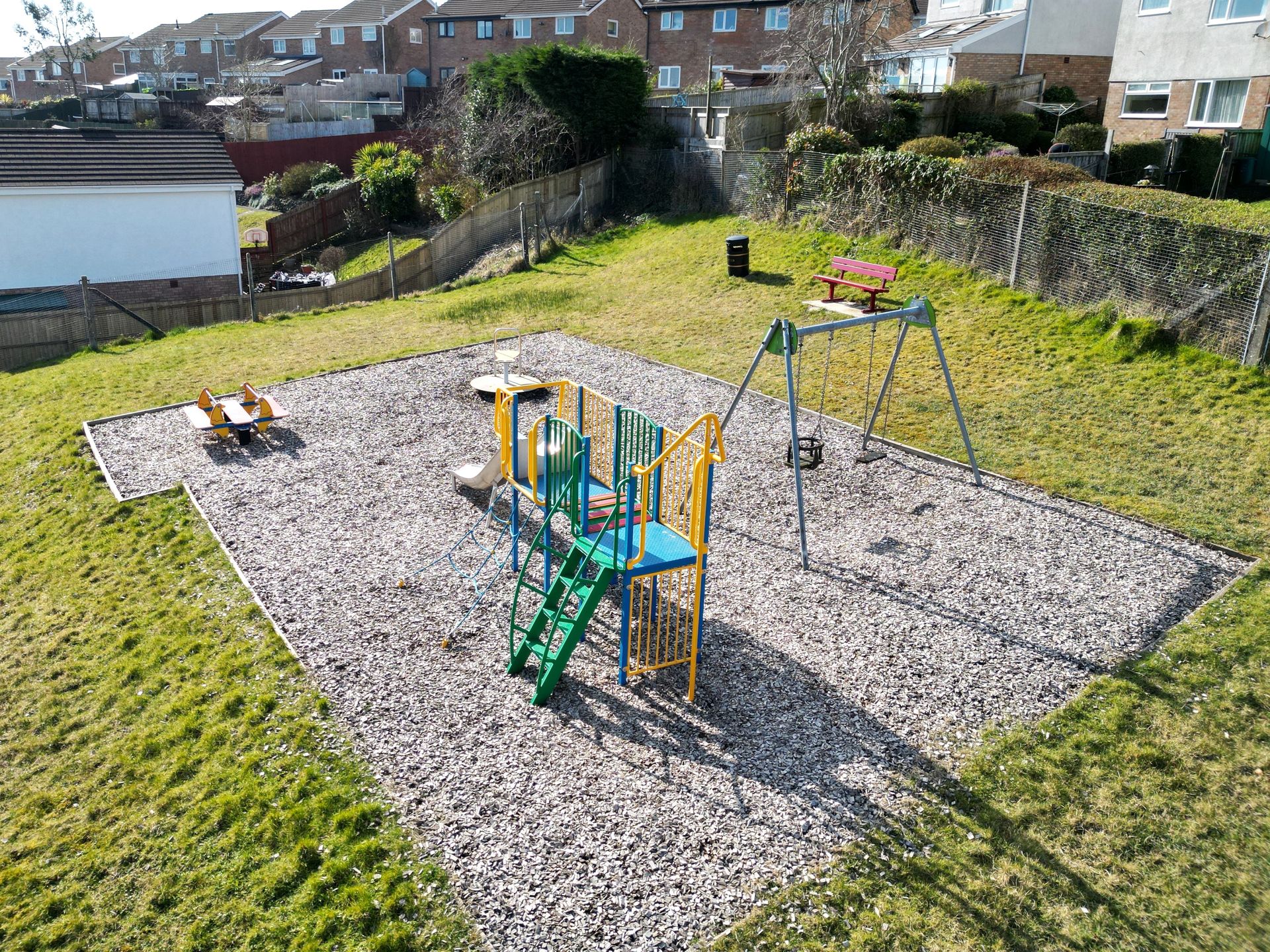 Playground with swing set, slides, and seesaw on a wood chip surface, surrounded by grass and fences.
