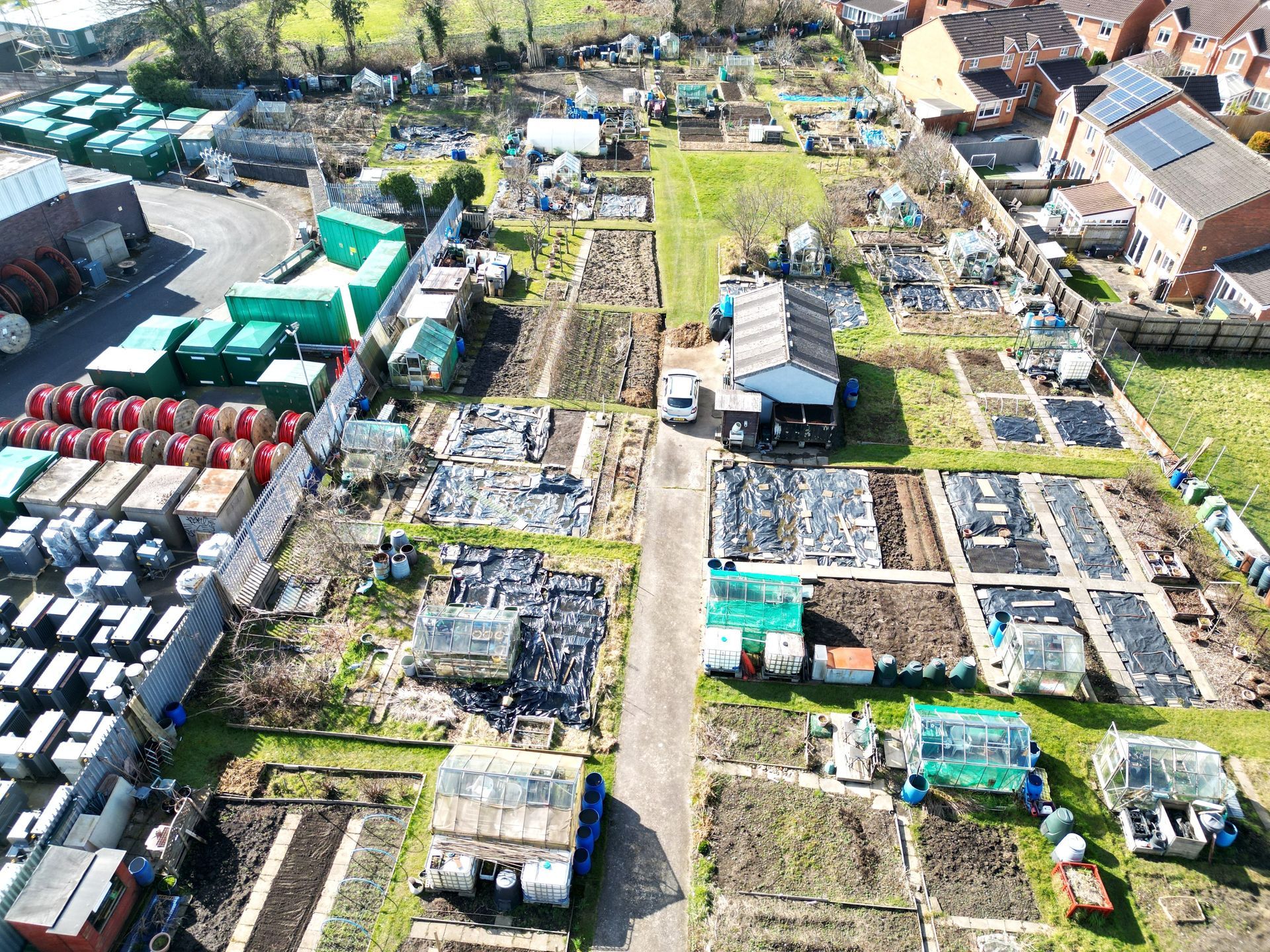 Aerial view of an allotment garden with plots, sheds, and equipment. Bright sunlight.