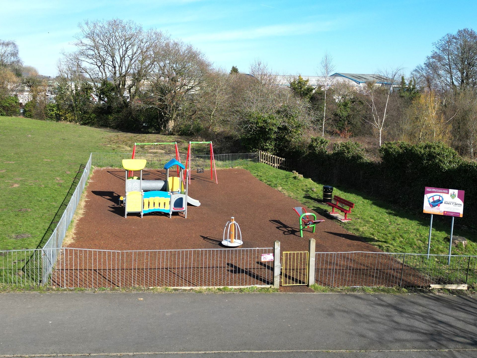 Playground with slides, swings, and other equipment on a rubberized surface, beside a grassy hill, under a blue sky.