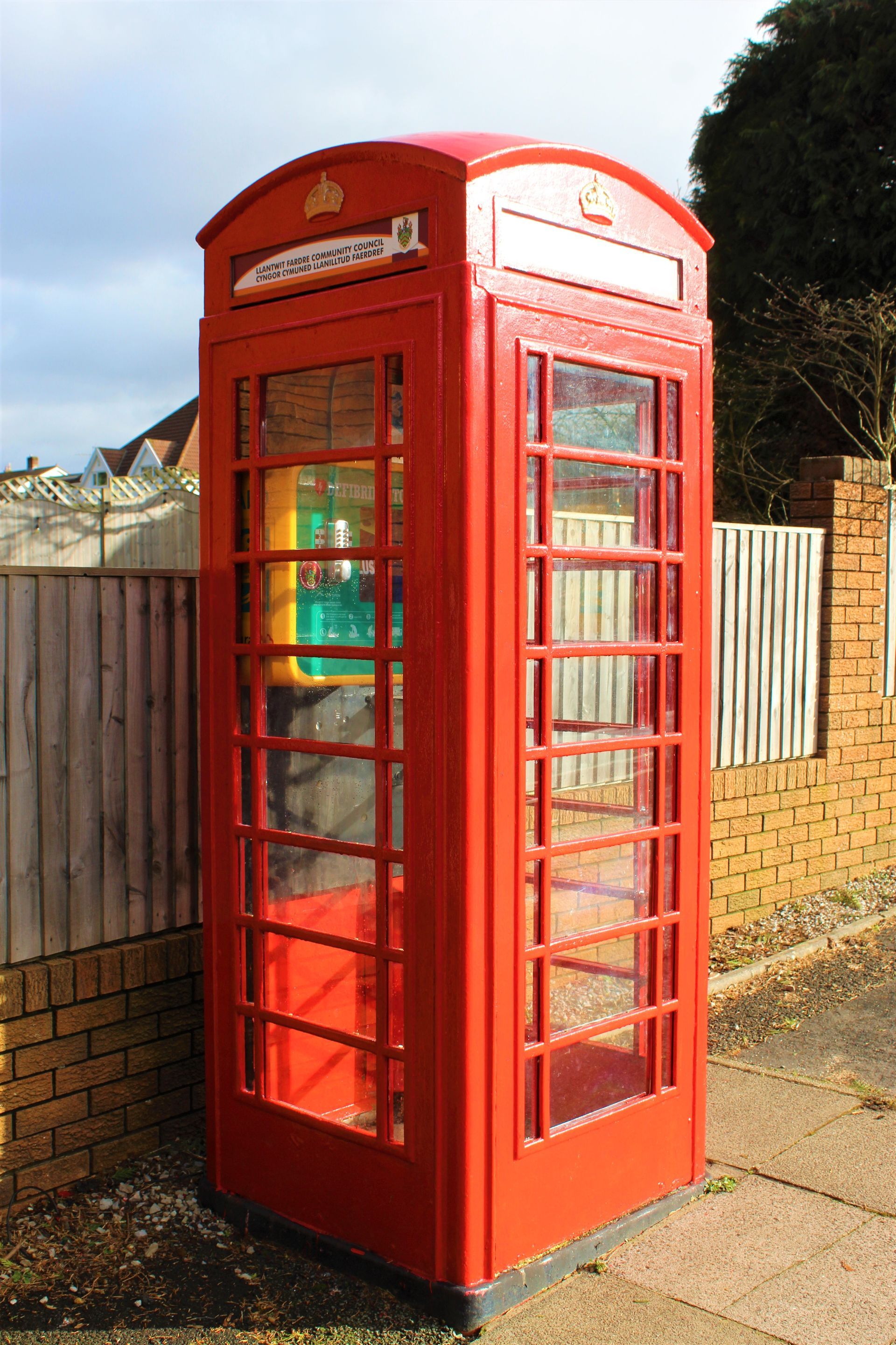 Red British telephone box on a sidewalk, glass windows, brick and wooden fence backdrop.