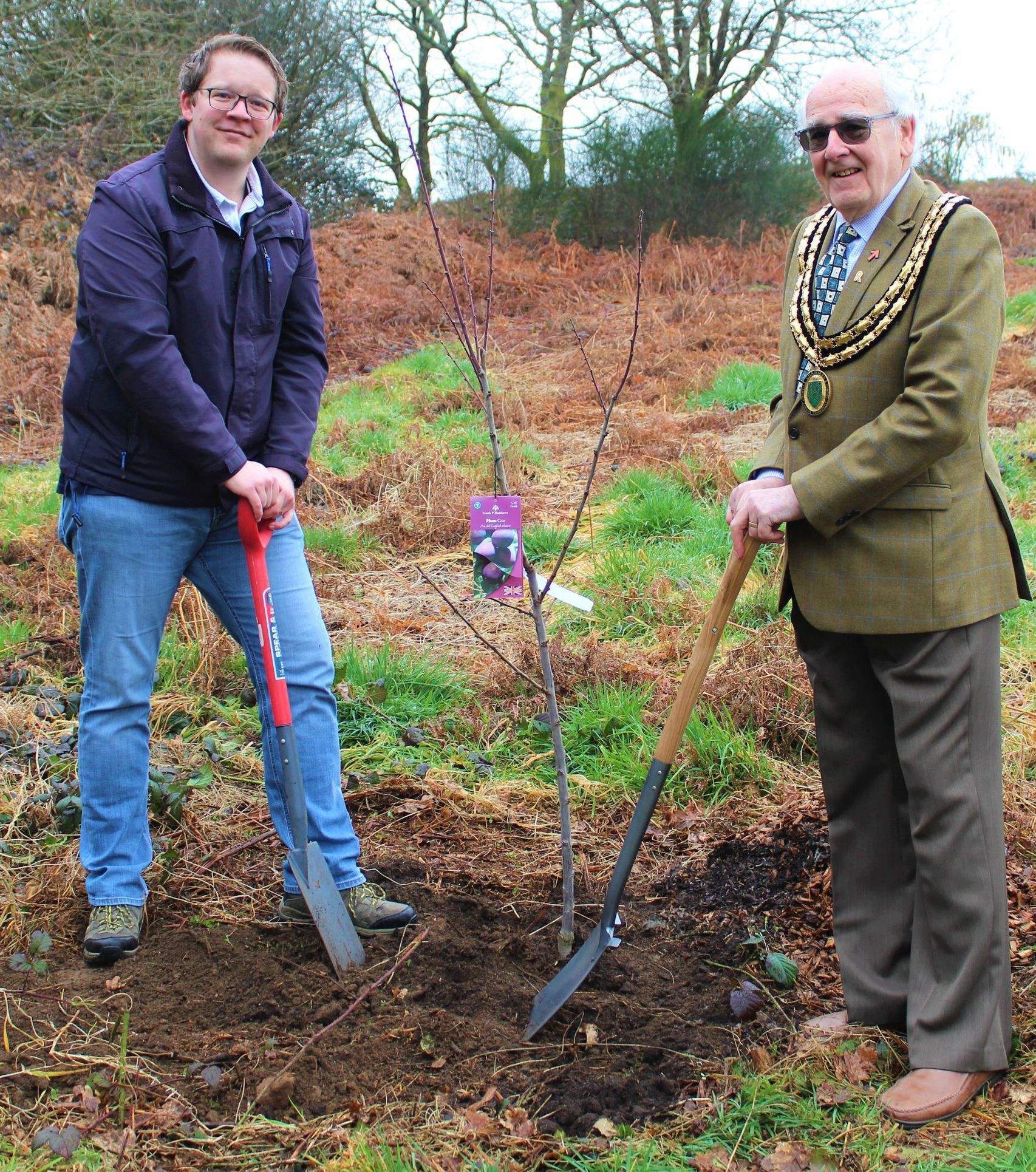 Two men planting a tree in an outdoor field. One man wears a jacket and jeans; the other, a suit and chain.